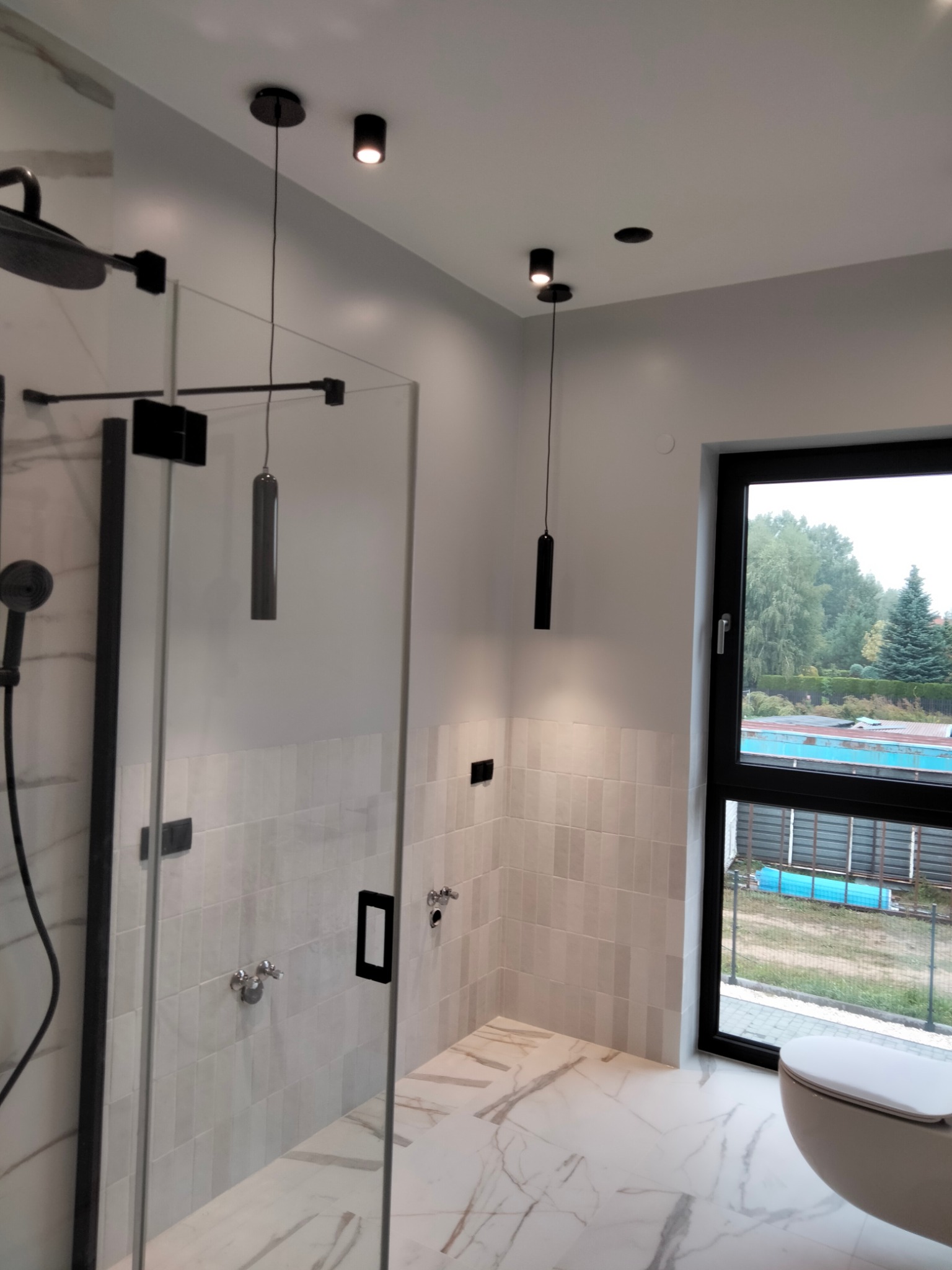 Nowoczesna łazienka z kabiną prysznicową typu walk-in, białymi płytkami imitującymi cegłę na ścianach, marmurową podłogą i czarnymi elementami wykończeniowymi oraz wiszącymi lampami.