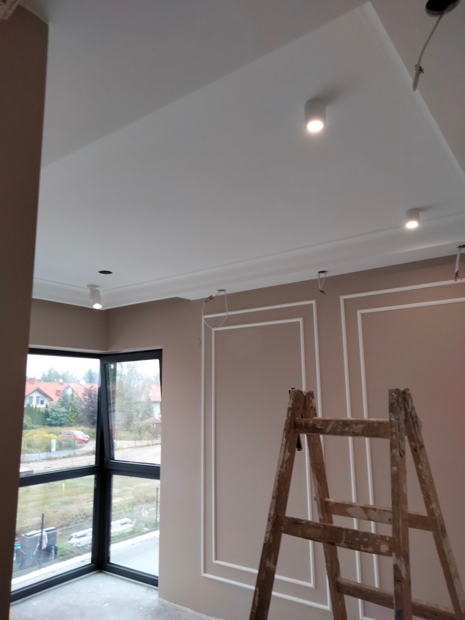 Wnętrze w Warszawie po remoncie, z pomalowanymi ścianami w odcieniach beżu, sztukaterią, białym sufitem z oświetleniem punktowym i drewnianą drabiną. Widok na zewnątrz przez okno.