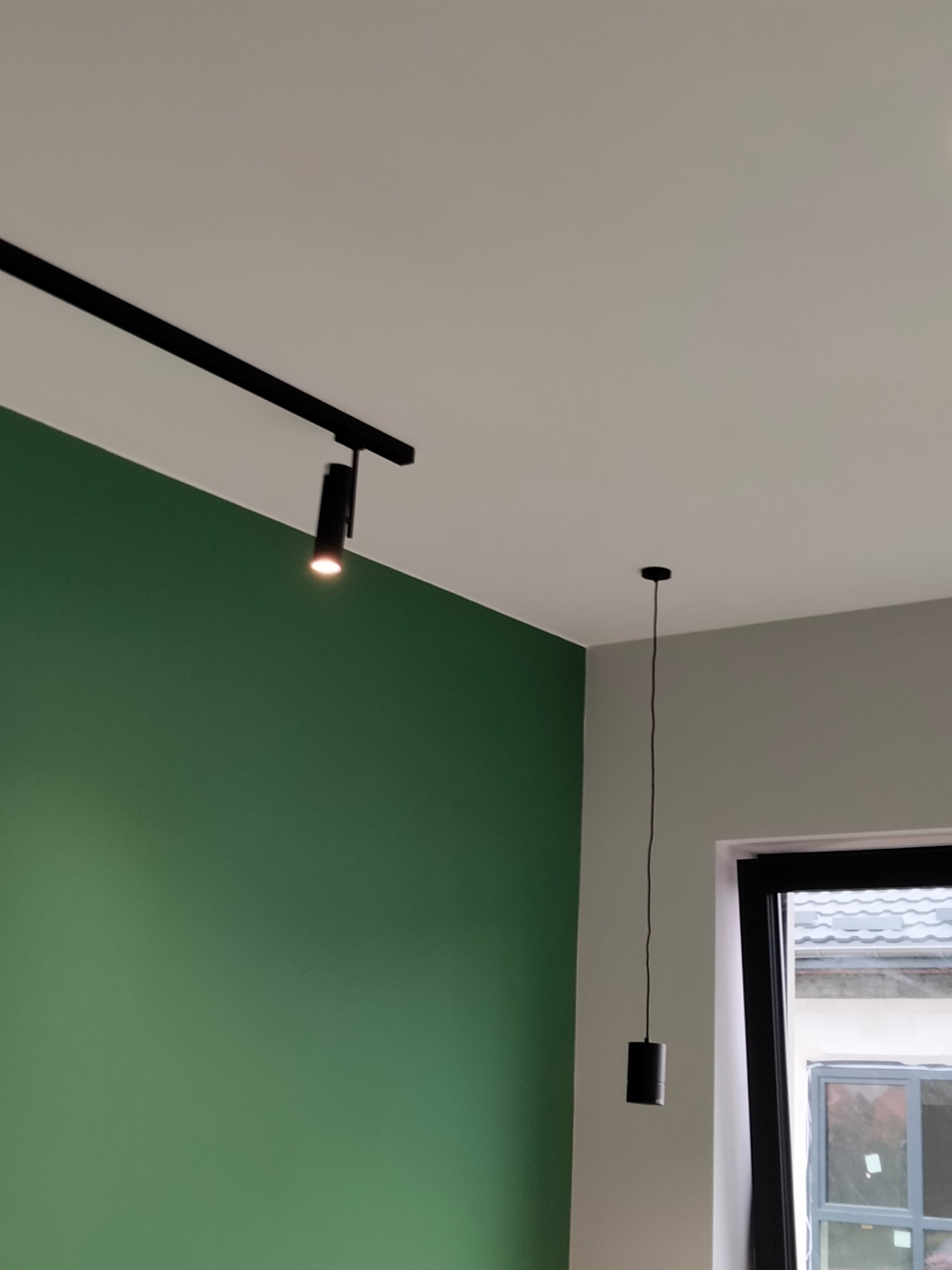 Ściana pomalowana na zielono z zamontowanym czarnym systemem szynowym oświetlenia punktowego oraz pojedyncza lampa wisząca na tle okna.