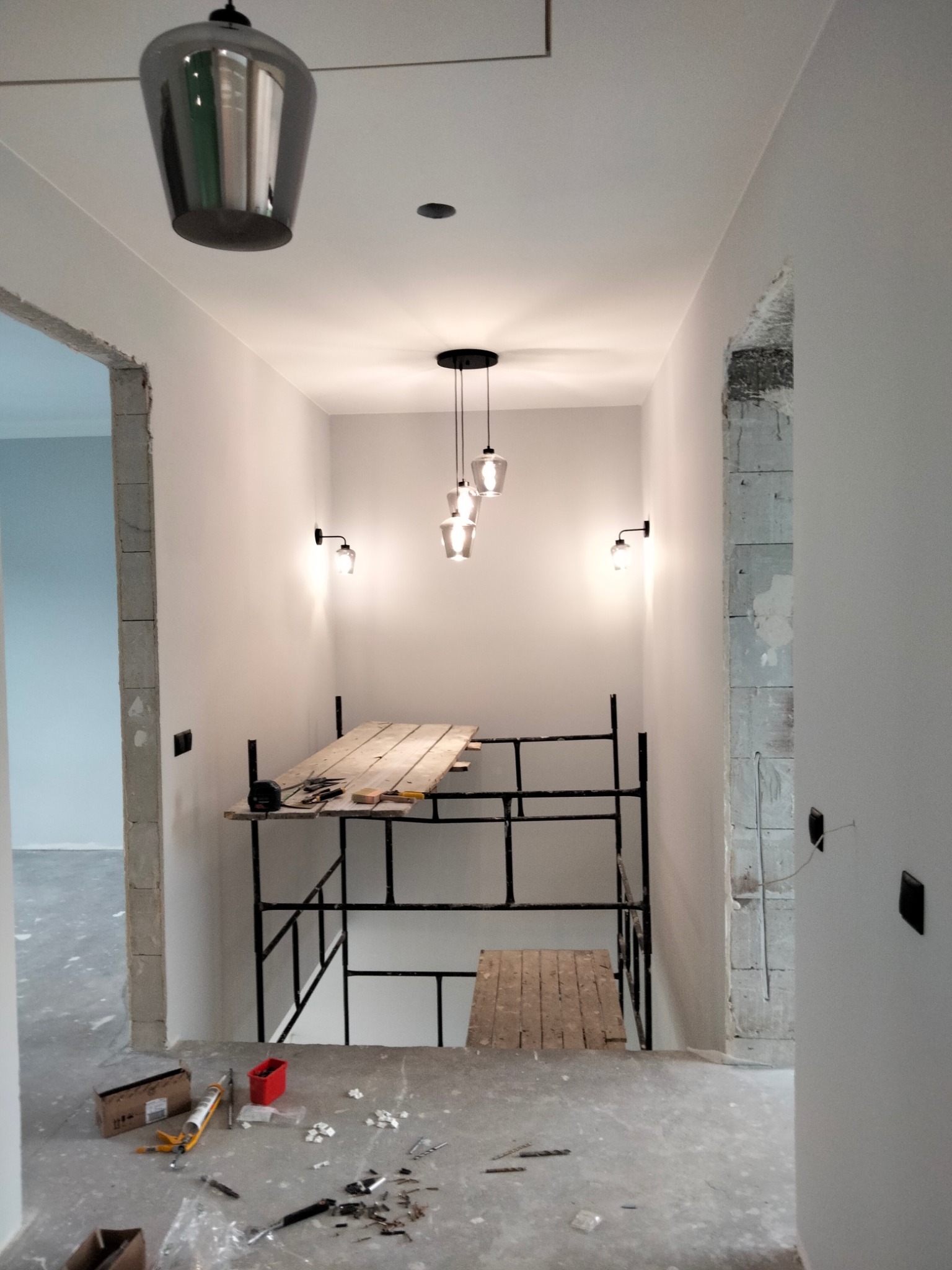 Wnętrze w trakcie remontu: białe ściany, surowy betonowy podkład, rusztowanie z drewnianym pomostem, narzędzia i akcesoria budowlane rozłożone na podłodze, wiszące lampy.