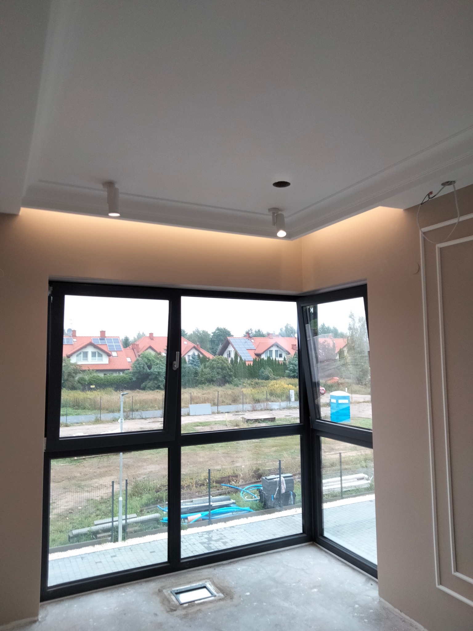 Wnętrze pokoju z dużym narożnym oknem z czarną ramą, widok na domy z panelami słonecznymi, sufit z oświetleniem ledowym i ozdobną sztukaterią, ściany w kolorze beżowym z dekoracyjnymi listwami.