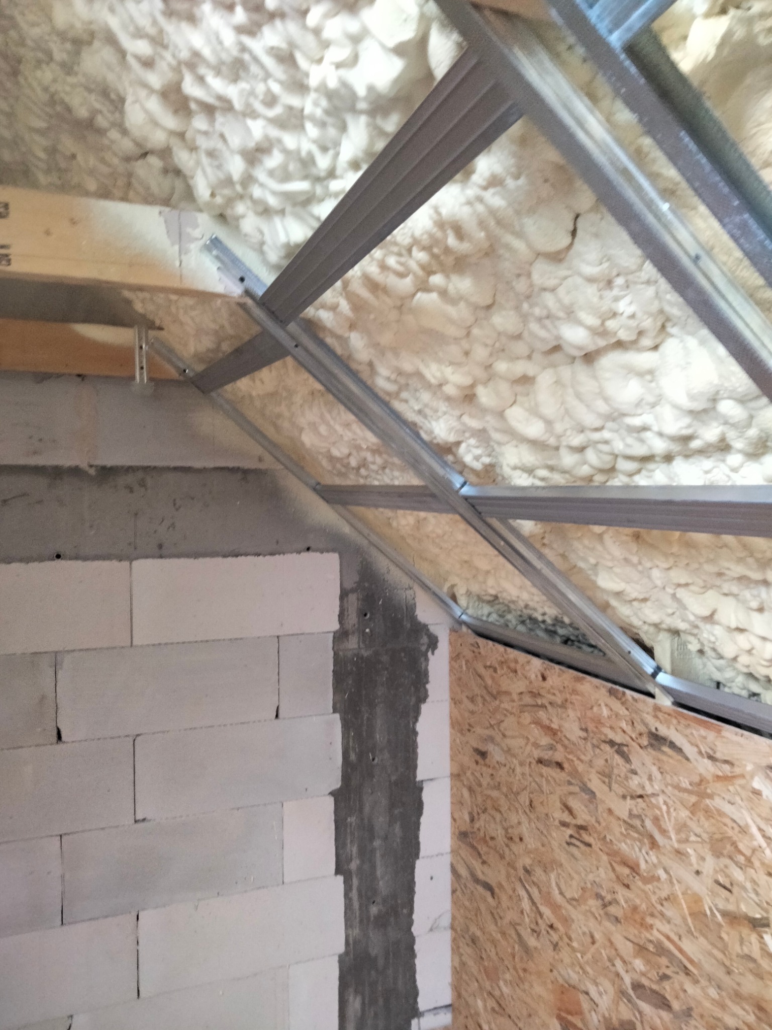 Poddasze w trakcie izolacji pianą natryskową, widoczne metalowe profile konstrukcyjne, ściana z betonu komórkowego i płyta OSB.