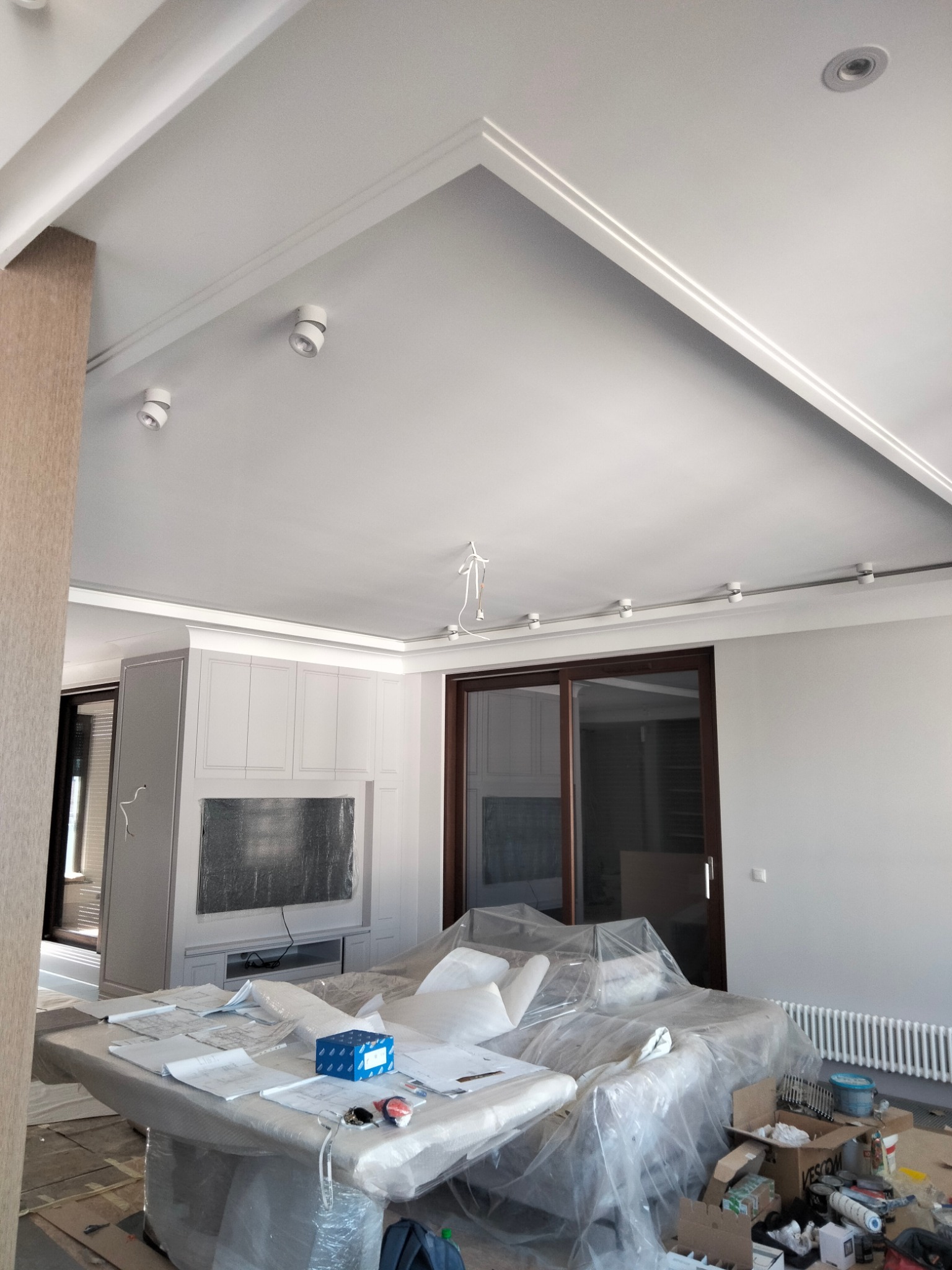 Wnętrze w trakcie remontu: zabezpieczone meble folią, widoczne prace wykończeniowe sufitu z listwami dekoracyjnymi i oświetleniem punktowym, na stole rozłożone plany i narzędzia.