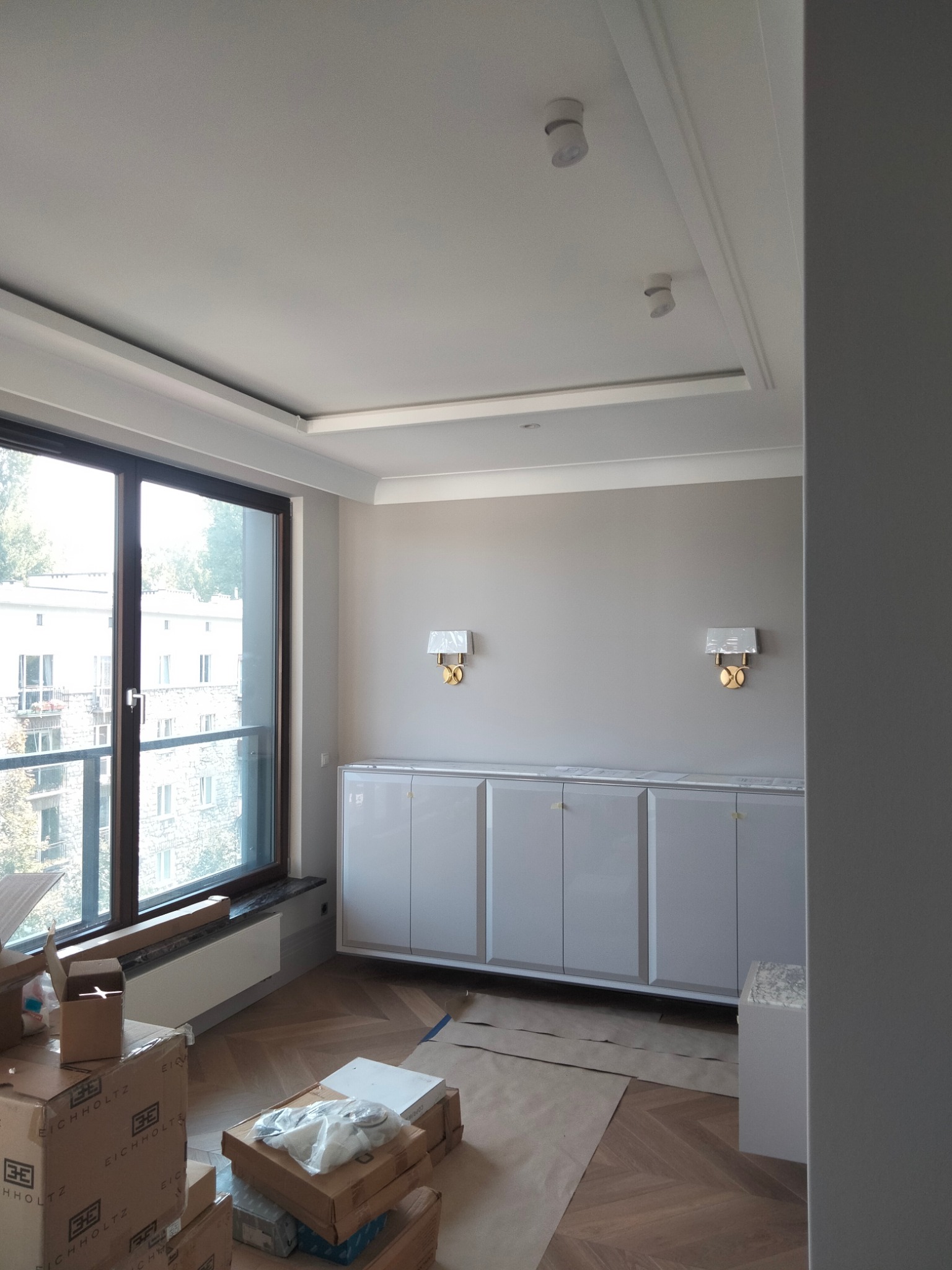 Jasne wnętrze po remoncie z parkietem w jodełkę, zabudową meblową pod ścianą, dużym oknem i kartonami po sprzętach na podłodze.