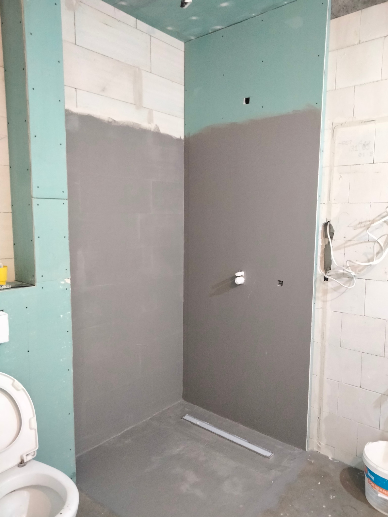 Niedokończona łazienka z szarymi ścianami, częściowo pokrytymi gładzią, widoczne płyty gipsowo-kartonowe, białe bloczki, otwarta toaleta i odpływ liniowy w podłodze.