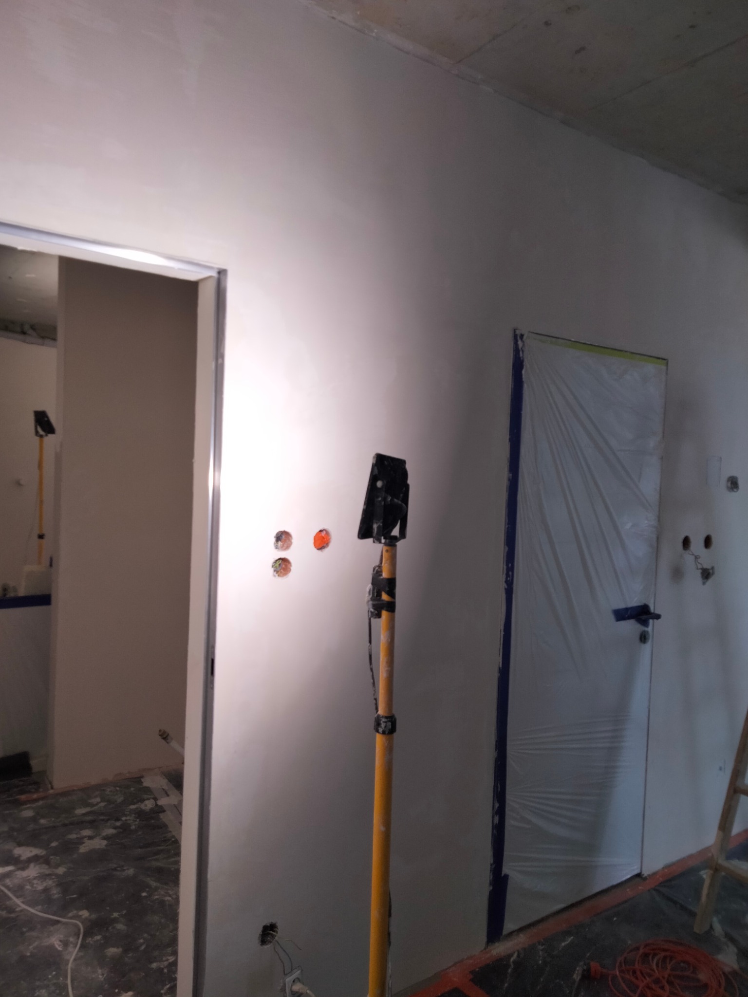 Wnętrze w trakcie prac wykończeniowych: ściana po nałożeniu gładzi, zabezpieczone folią drzwi, widoczne puszki elektryczne i lampa robocza na statywie.
