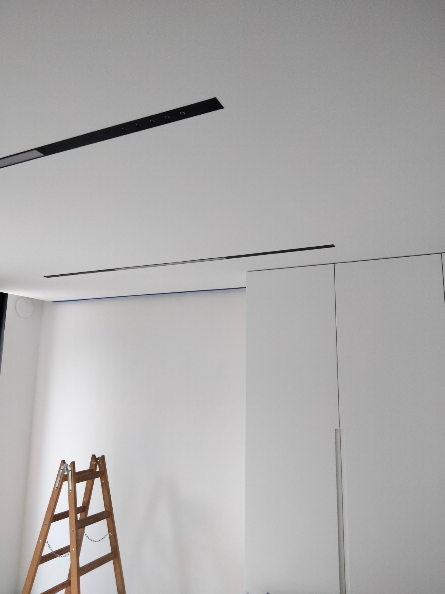 Wnętrze w trakcie prac wykończeniowych: drewniana drabina oparta o białą ścianę z widoczną taśmą malarską pod sufitem, minimalistyczne czarne lampy sufitowe i biała szafa w tle.