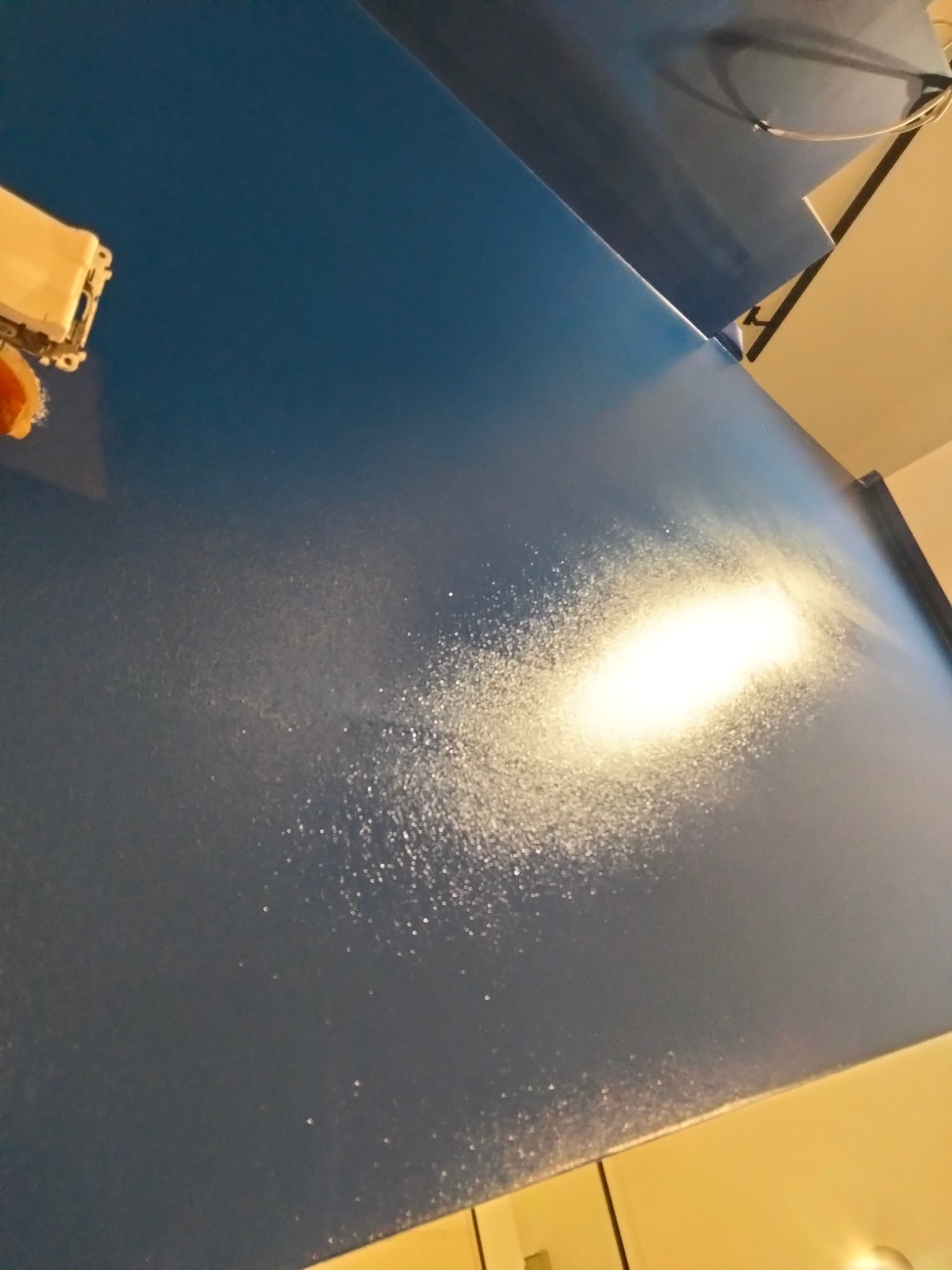 Niebieska ściana po szpachlowaniu, widoczne drobne nierówności i pył, przygotowana do malowania, z widoczną puszką elektryczną w rogu.