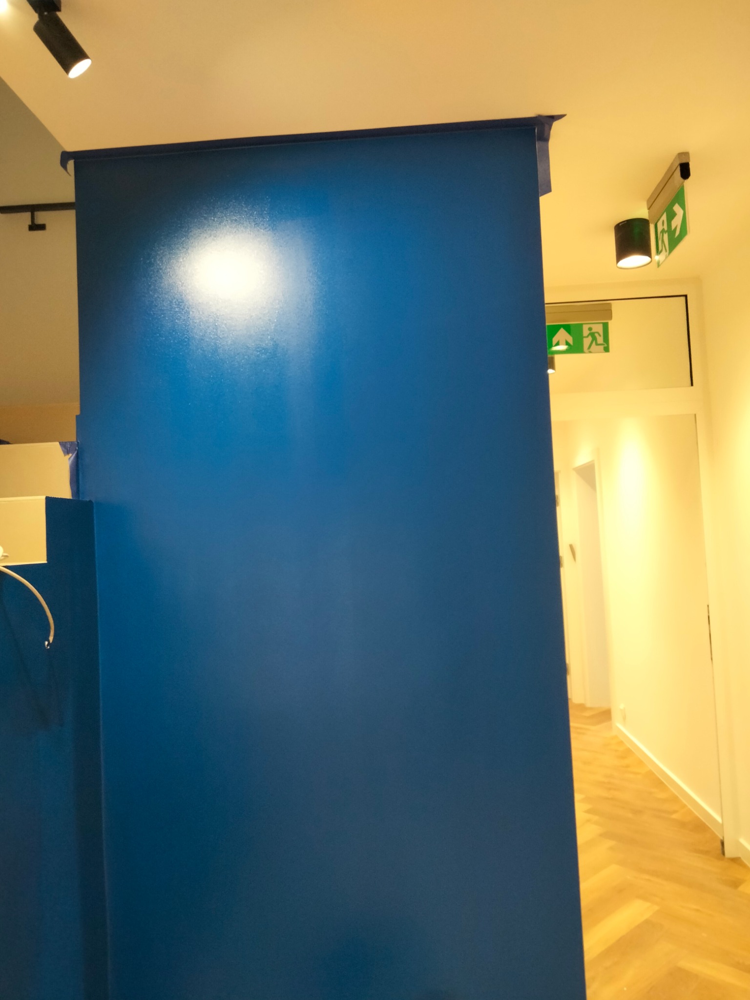 Świeżo pomalowana na niebiesko ściana z taśmą malarską zabezpieczającą krawędzie, widoczny fragment podłogi z parkietem w jodełkę i oświetlenie sufitowe z oznaczeniem drogi ewakuacyjnej.
