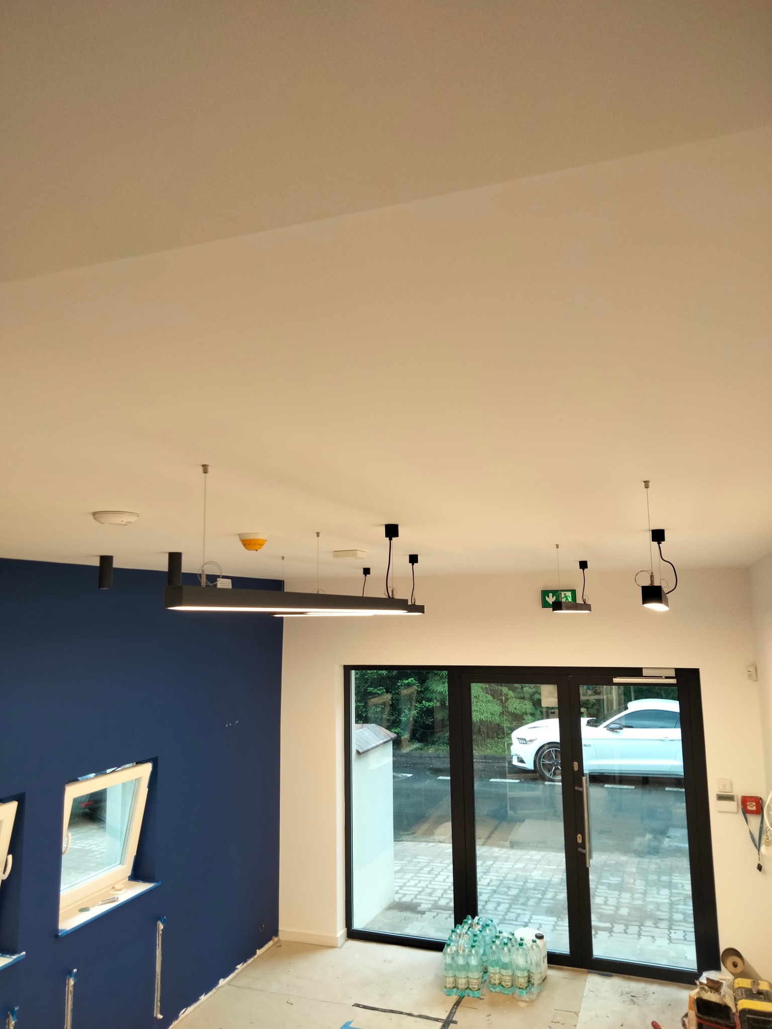 Wnętrze w trakcie prac wykończeniowych: pomalowana na granatowo ściana z oknami, biały sufit z nowoczesnym oświetleniem, podłoga częściowo zakryta folią, widoczne butelki wody i narzędzia.
