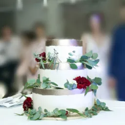 Trzypiętrowy tort weselny z białym kremem, złotymi akcentami i dekoracją z ciemnoczerwonych goździków oraz eukaliptusa, na tle rozmytych postaci gości.