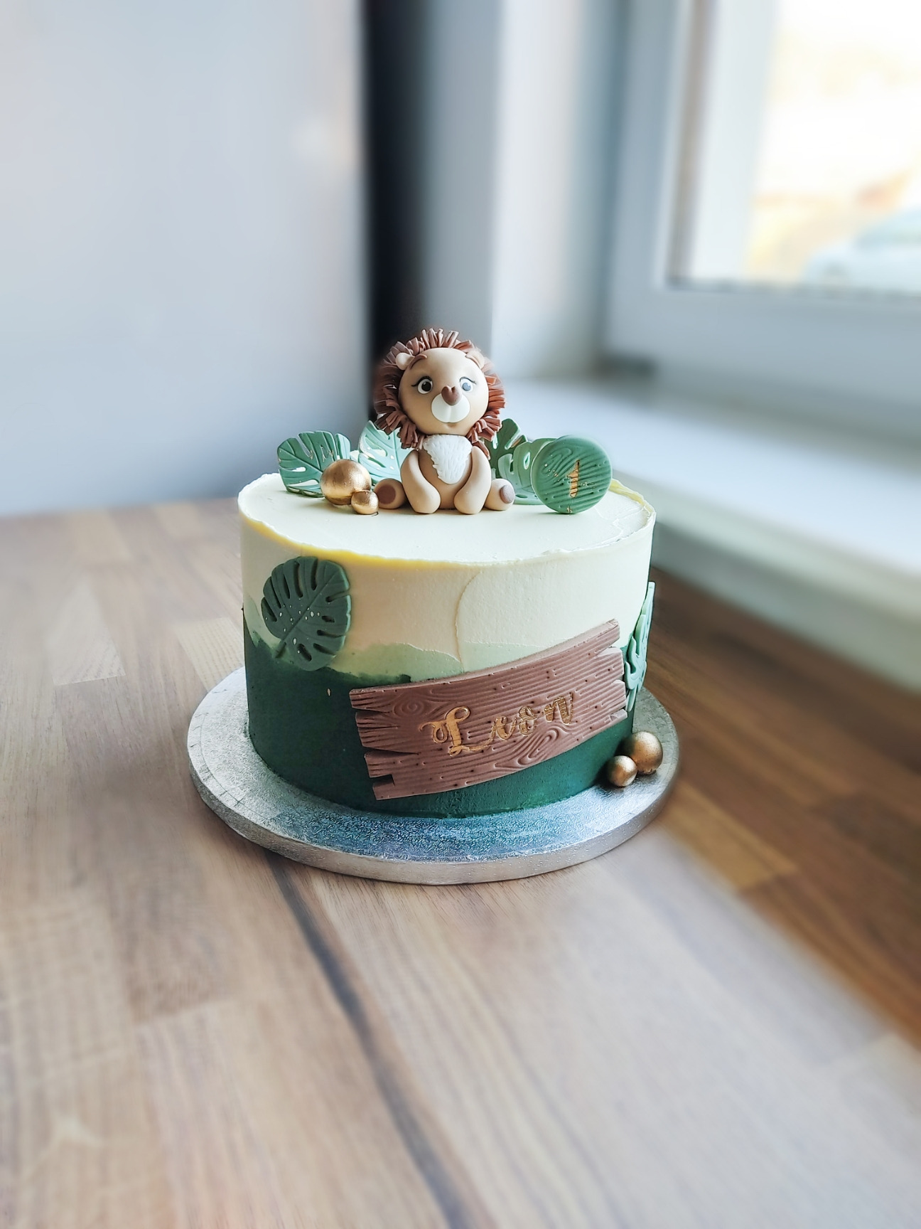Tort urodzinowy z motywem dżungli, dekorowany figurką lwa z lukru, zielonymi liśćmi i napisem na drewnianej tabliczce, umieszczony na srebrnej paterze na drewnianym stole.