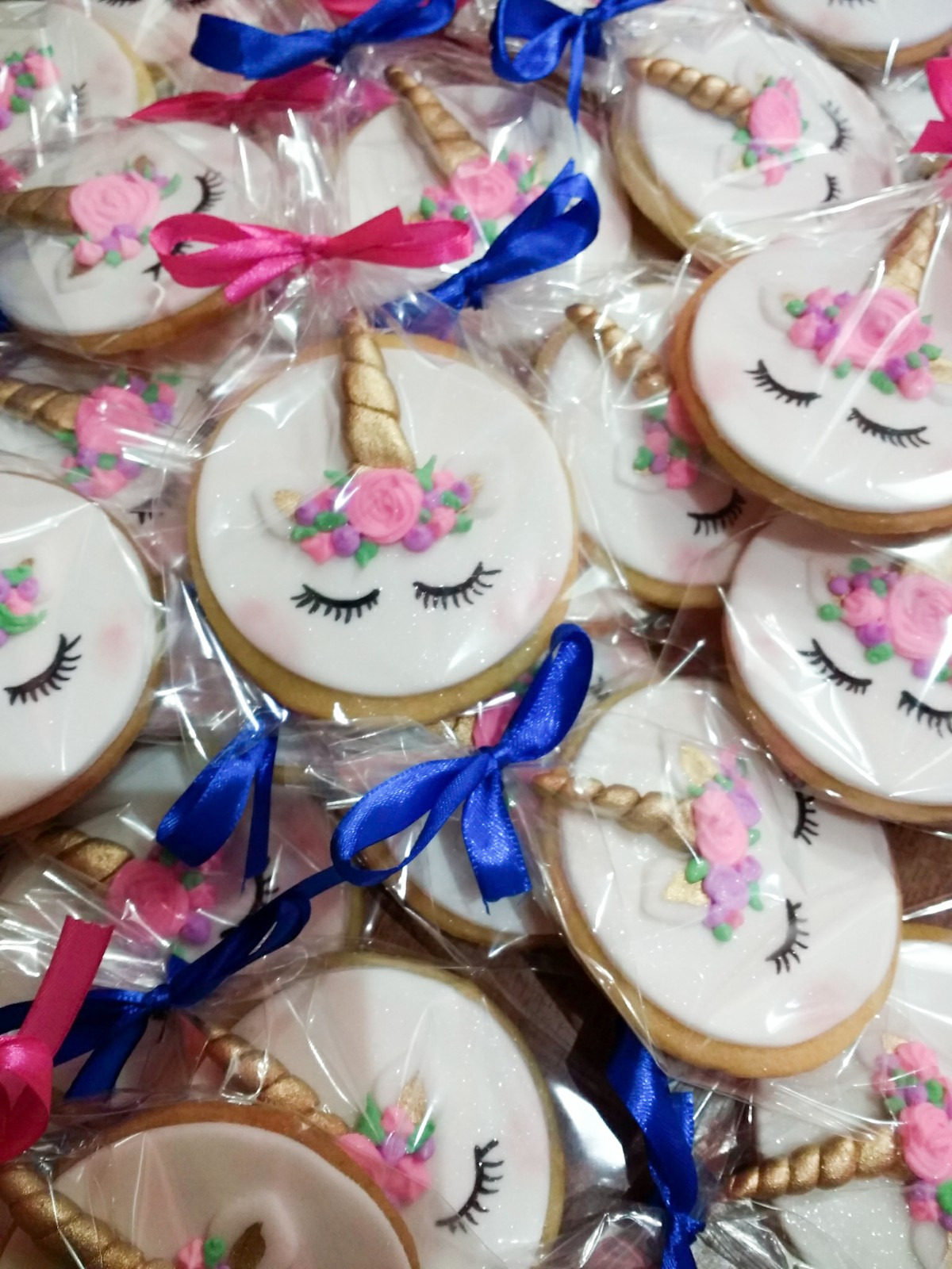 Ręcznie robione, okrągłe ciasteczka z lukrem, dekorowane motywem jednorożca z zamkniętymi oczami, kwiatami i złotym rogiem, zapakowane w przezroczyste folie i przewiązane różowymi i niebieskimi...