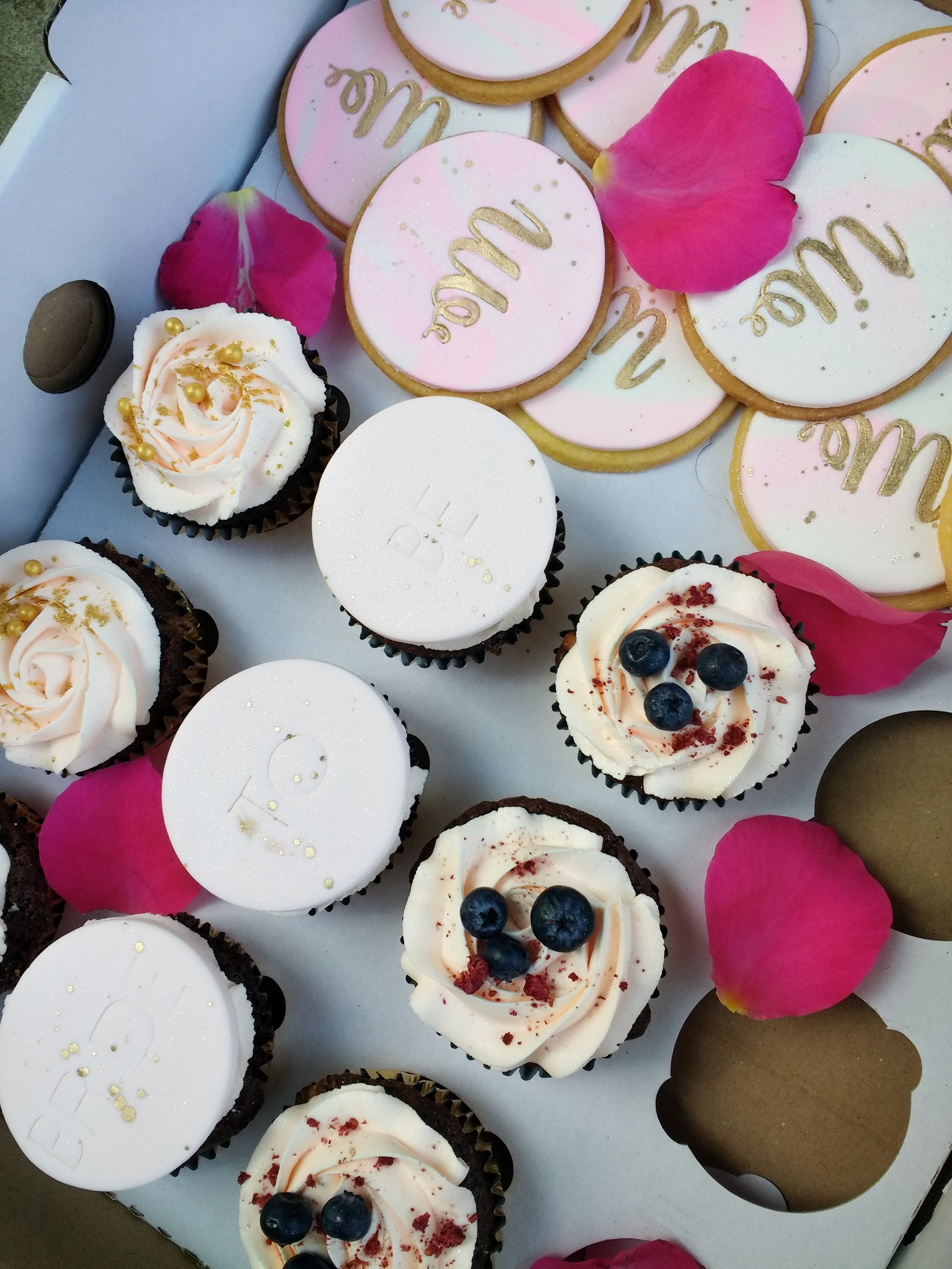 Kolekcja babeczek i ciasteczek w pudełku, dekorowanych kremem, owocami i złotymi akcentami, otoczona płatkami róż.