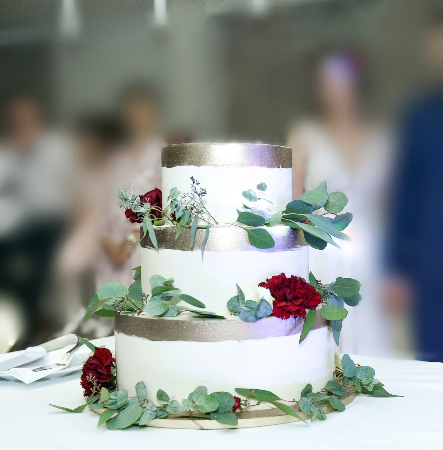 Trzypiętrowy tort weselny z białym kremem, złotymi akcentami i dekoracją z ciemnoczerwonych goździków oraz eukaliptusa, na tle rozmytych postaci gości.