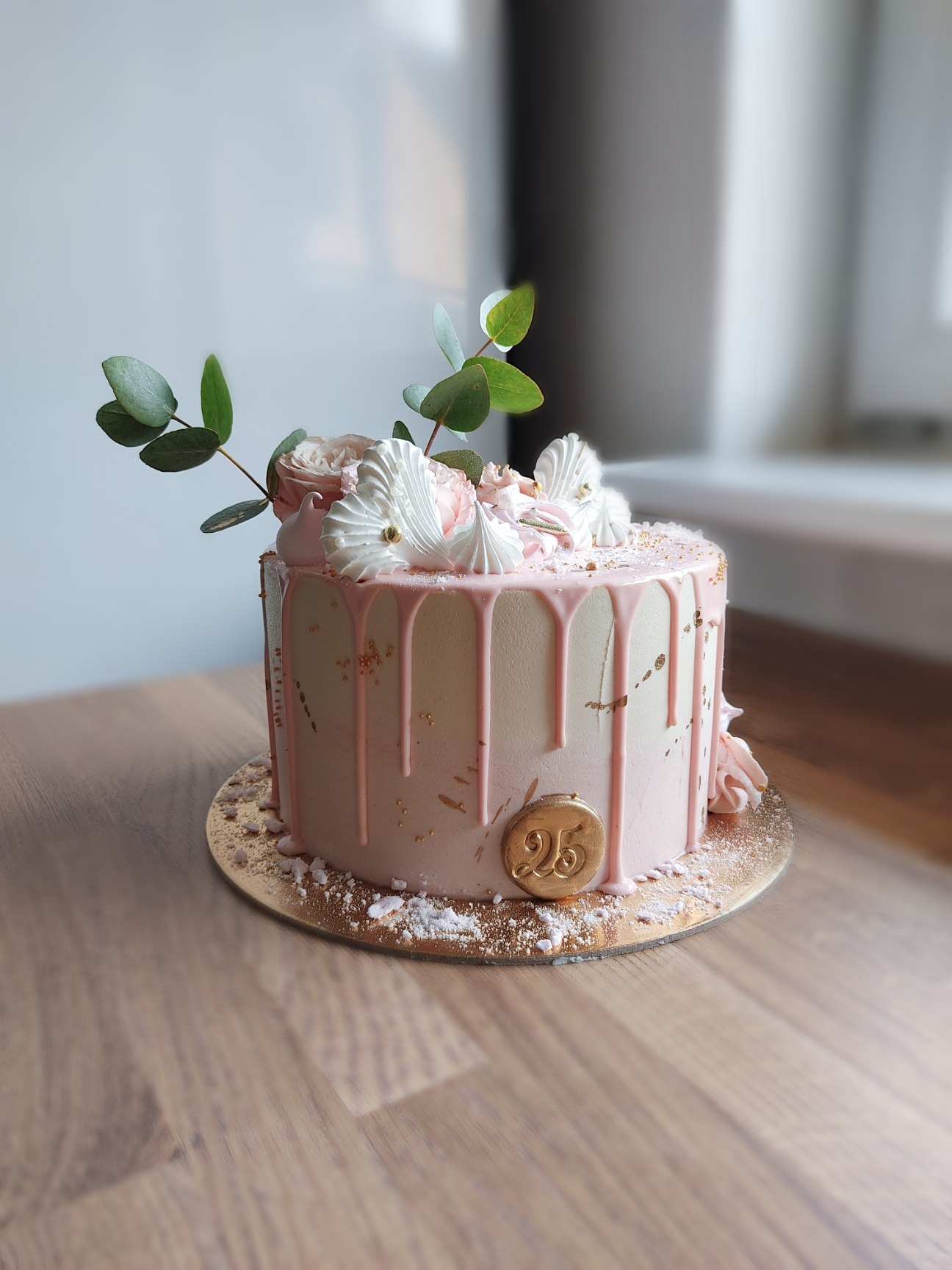 Tort urodzinowy w pastelowych barwach z różowym lukrem spływającym po bokach, udekorowany białymi bezami, różami i gałązkami eukaliptusa, z okrągłą złotą dekoracją z liczbą 25.