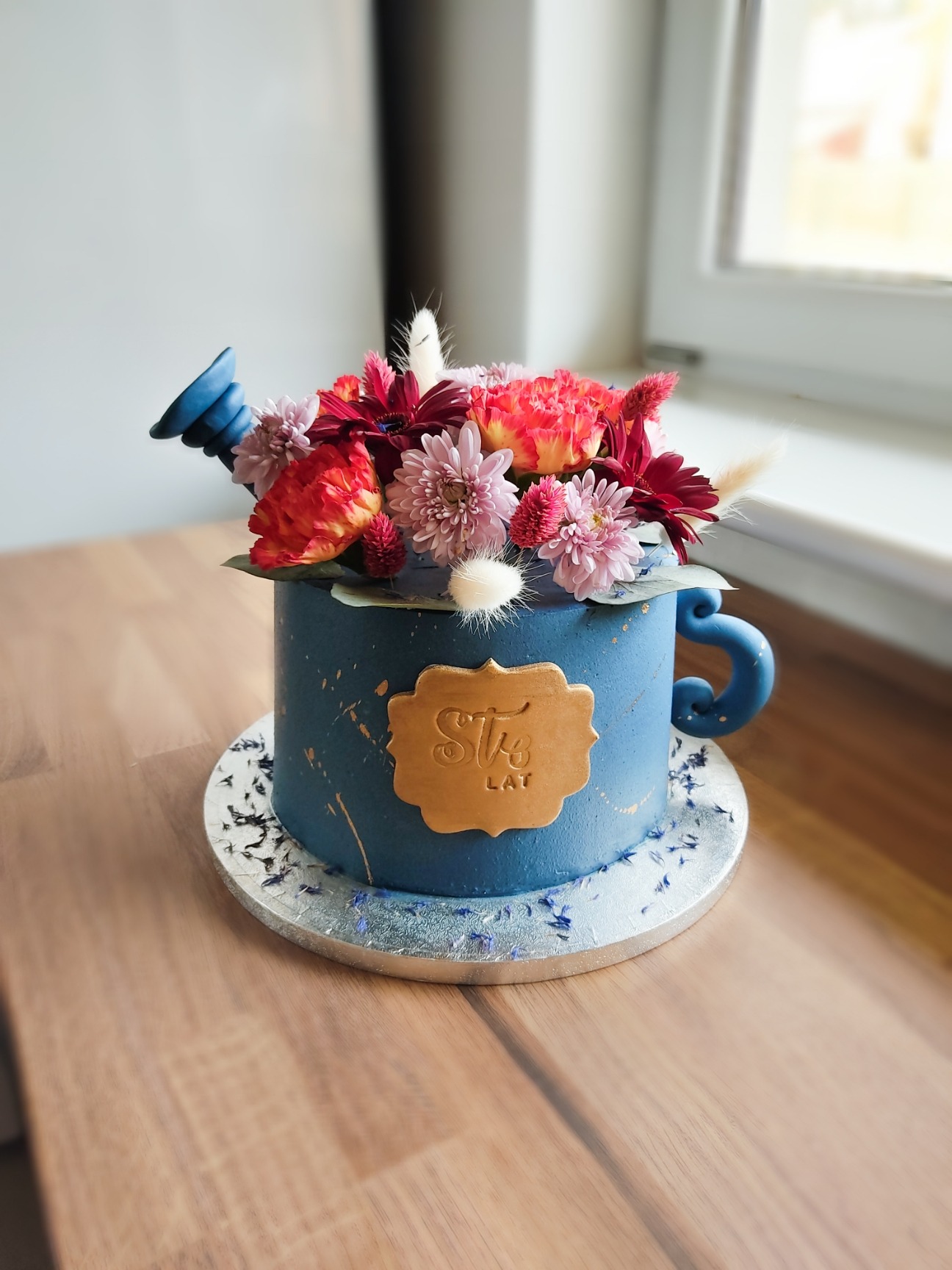 Tort w kształcie niebieskiego kubka z uchwytem i wylewką, udekorowany świeżymi kwiatami i złotą tabliczką z napisem 'Sto lat', umieszczony na srebrnej podkładce.