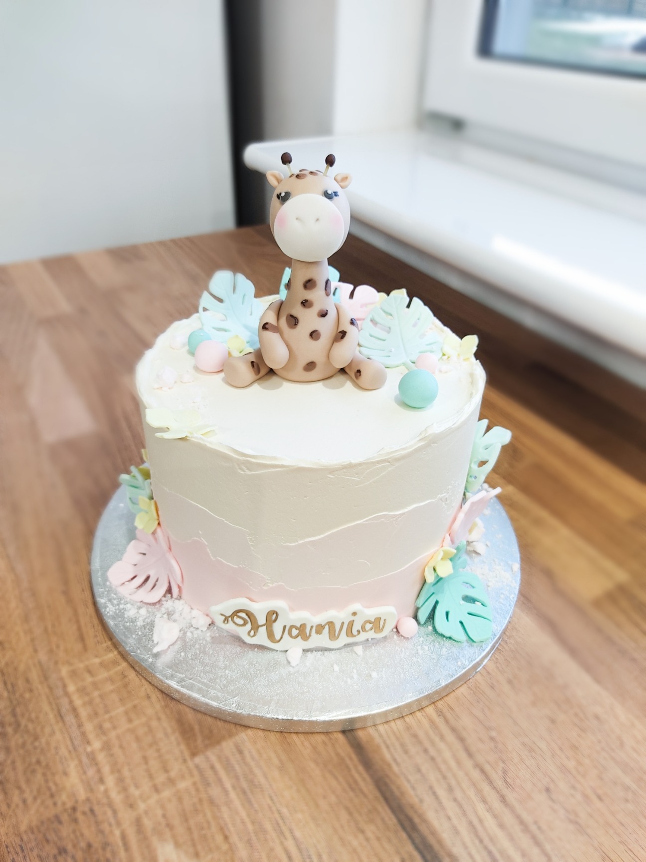 Tort urodzinowy z motywem żyrafy, pastelowe kolory, dekoracje z liści i imię 'Hania' na froncie.