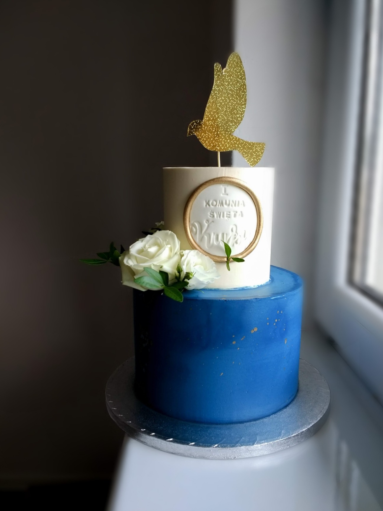 Dwupoziomowy tort w kolorze granatowym i białym, dekorowany białymi różami i złotym gołębiem, z napisem 'I Komunia Święta'.