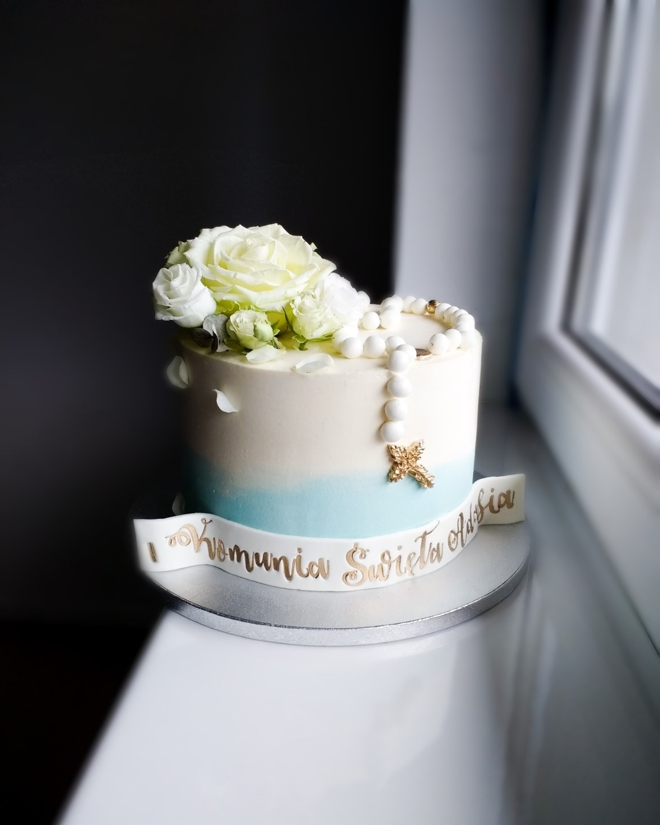 Tort komunijny z białymi różami i różańcem, dekorowany ombre w błękicie i napisem 'I Komunia Święta Adasia'.
