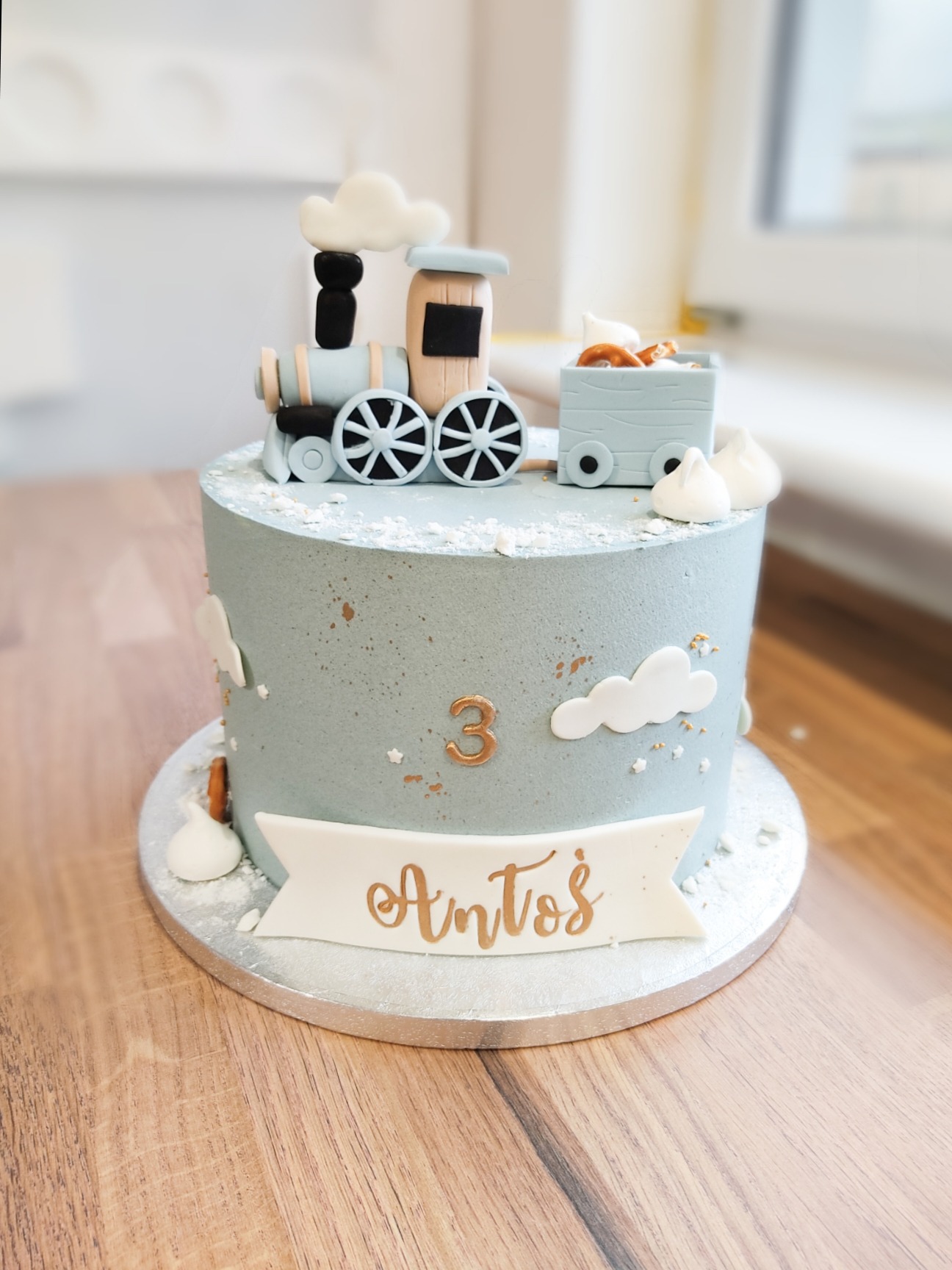 Tort urodzinowy w pastelowych odcieniach z dekoracją w kształcie pociągu i napisem 'Antoś' na białym banerze, cyfra '3' z boku.