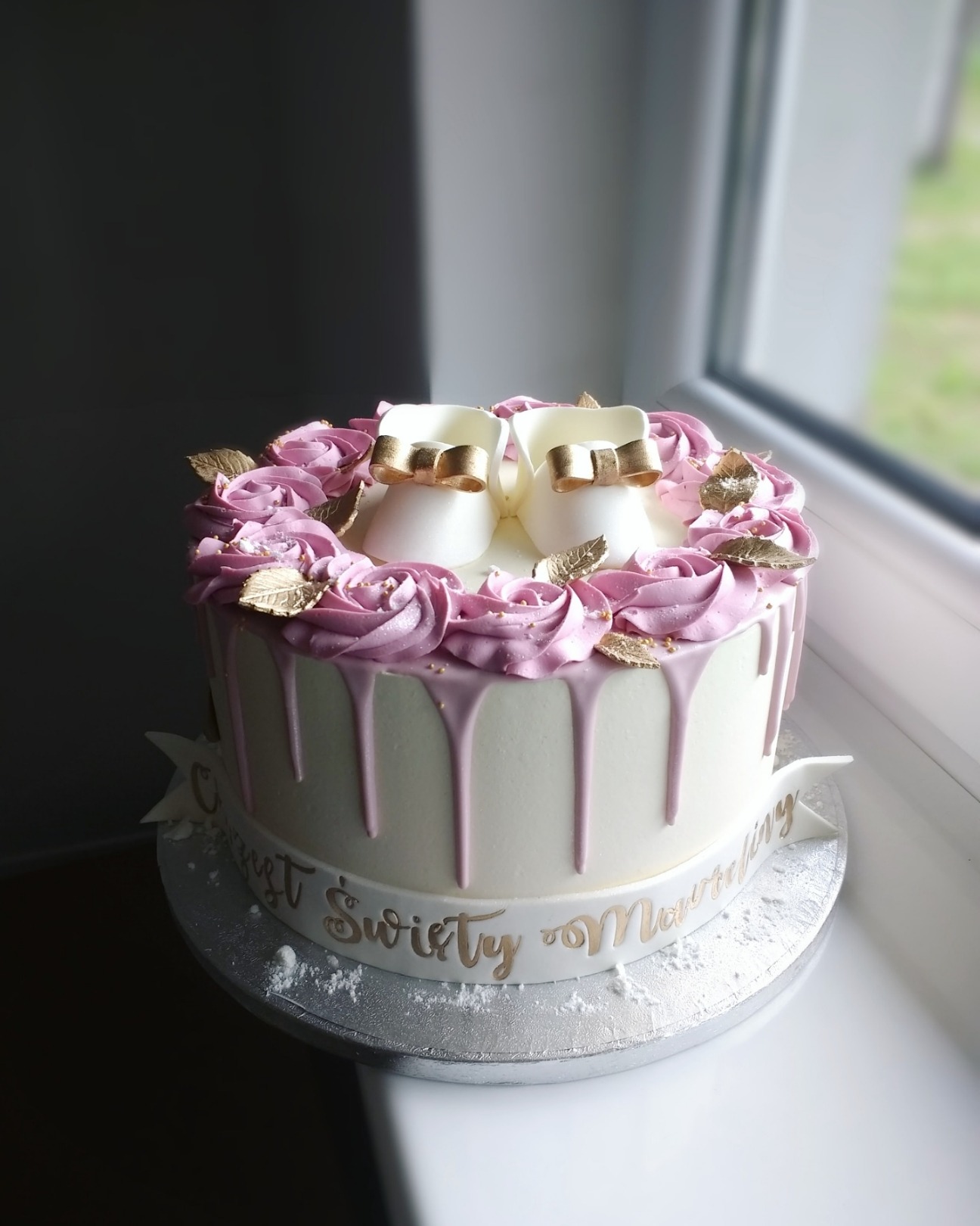 Tort na Chrzest Święty z dekoracją z różowych róż, złotych listków i białych bucików z kokardkami, na srebrnej paterze przy oknie.