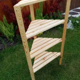 Tr&oacute;jkątna, drewniana p&oacute;łka na rośliny, wykonana z jasnego drewna, ustawiona na trawniku w Brz&oacute;zie Stadnickiej, z widocznymi roślinami w tle.