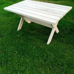 Drewniany stolik ogrodowy z jasnego drewna, ustawiony na zielonej trawie, z widocznymi paletami w tle.