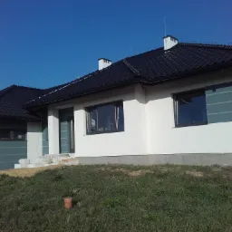 Nowoczesny dom jednorodzinny z białą elewacją i szarymi akcentami, czarną dachówką, widoczne elementy wykończeniowe i niedokończone schody wejściowe.