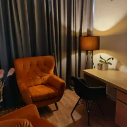 Pomarańczowy fotel Chesterfield w gabinecie z drewnianym biurkiem, lampą na trójnogu i czarnym krzesłem na drewnianej podłodze, zasłonięte okno w tle.