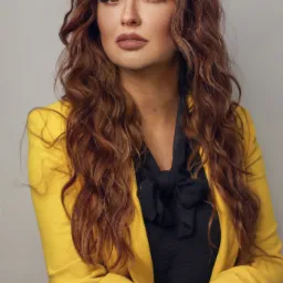 Portret kobiety z długimi, kręconymi, brązowymi włosami, ubrana w żółty żakiet i czarną bluzkę z kokardą, spoglądającej w dal.