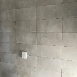 Ściana wyłożona szarymi, prostokątnymi płytkami ceramicznymi z widoczną puszką instalacyjną Grohe po lewej stronie, w rogu ściany widoczne zabezpieczenie z niebieskiej taśmy malarskiej.