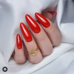 Dłoń z długimi, intensywnie czerwonymi paznokciami w kształcie migdała, ozdobiona złotym pierścionkiem z węzłem na serdecznym palcu, na tle białej, satynowej tkaniny.