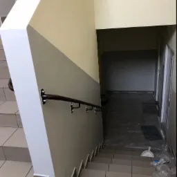 Klatka schodowa w trakcie prac wykończeniowych: pomalowane ściany w dwóch odcieniach, zabezpieczone folią podłogi i schody, widoczna poręcz i akcesoria do sprzątania.