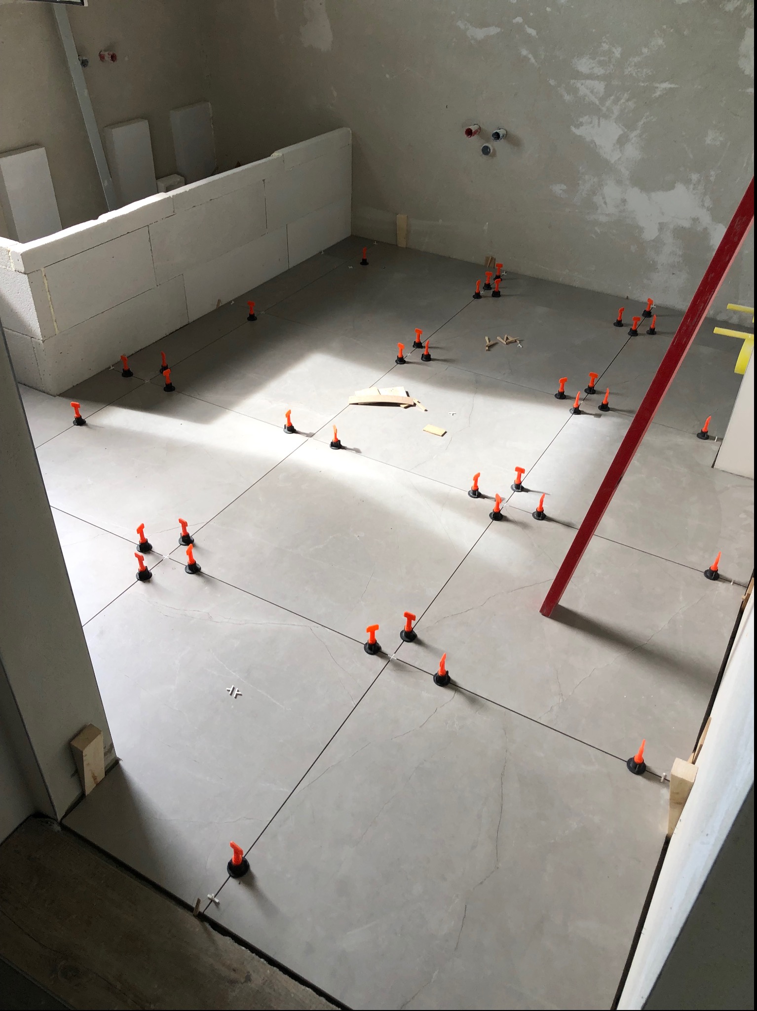 Szare płytki podłogowe w trakcie układania, z pomarańczowymi klipsami poziomującymi i czarnymi krzyżykami dystansowymi, widoczne linie traserskie, fragment ściany z surowego betonu i bloczków.