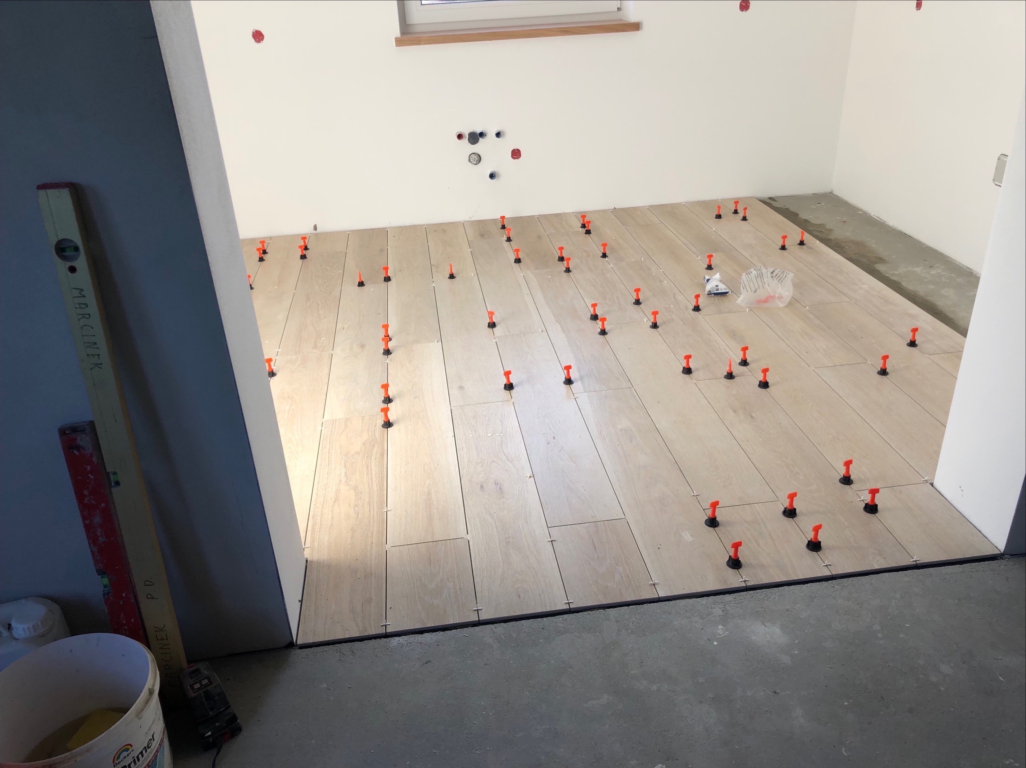 Układanie podłogi z paneli imitujących drewno, z użyciem poziomujących klipsów i klinów; widoczny poziomica i wiadro z gruntem.