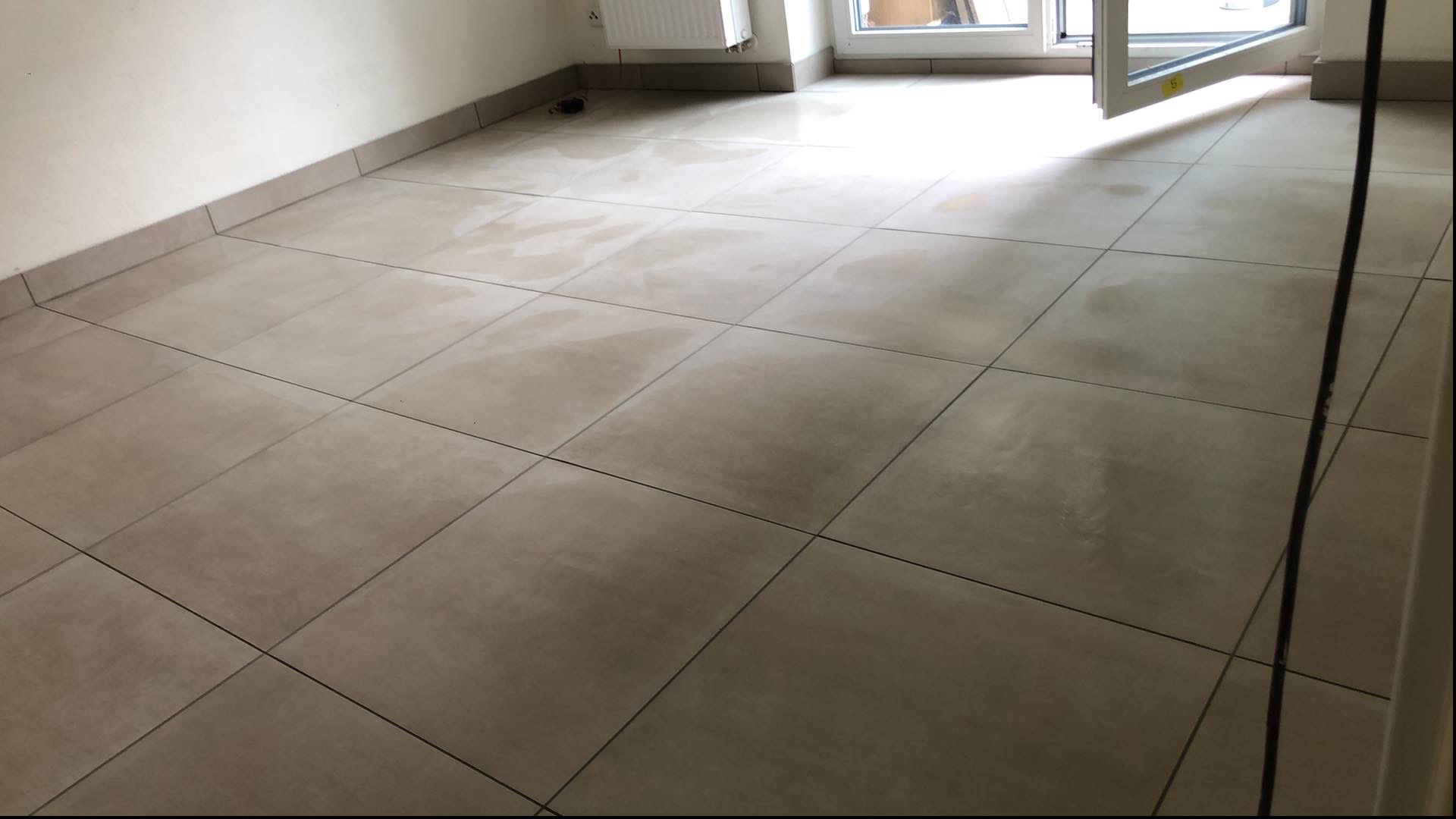 Beżowe, kwadratowe płytki podłogowe, świeżo po umyciu, z widocznymi smugami wilgoci, ułożone w pustym pomieszczeniu z białymi ścianami i otwartym balkonem.