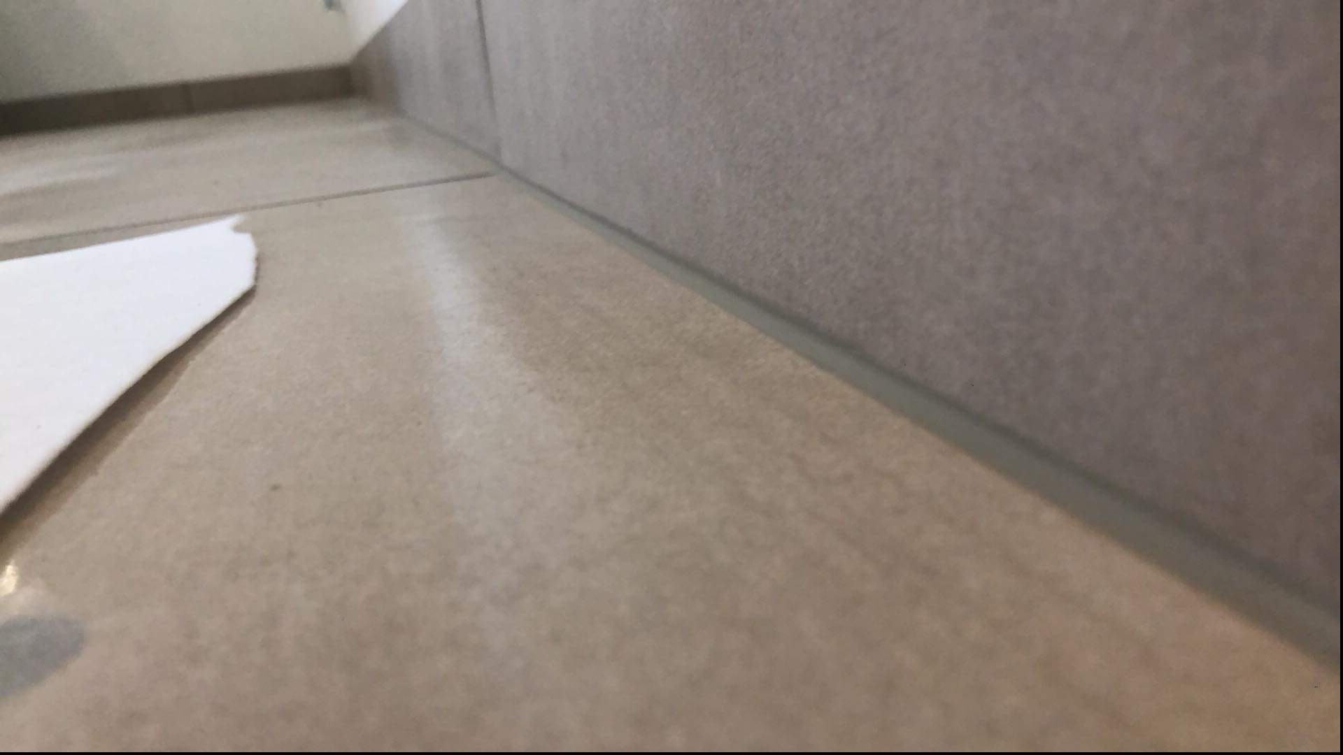 Beżowe płytki podłogowe i ścienne, połączone szarą fugą silikonową, z fragmentem białego papieru zabezpieczającego, ujęcie z niskiej perspektywy.