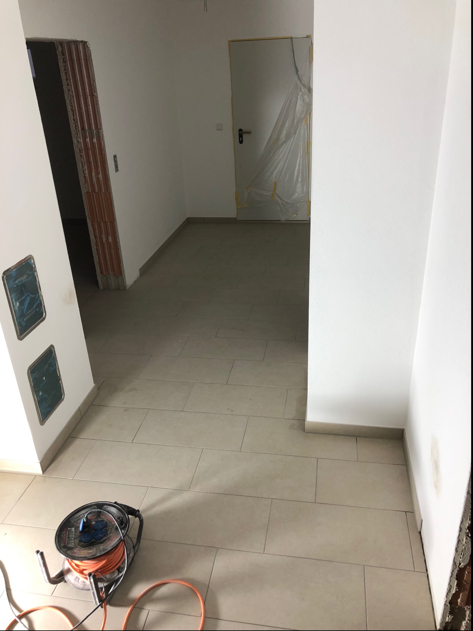 Wnętrze w trakcie prac wykończeniowych: świeżo pomalowane białe ściany, ułożona jasna podłoga z płytek, zabezpieczone gniazdka elektryczne i drzwi folią malarską, widoczny fragment surowej cegły...