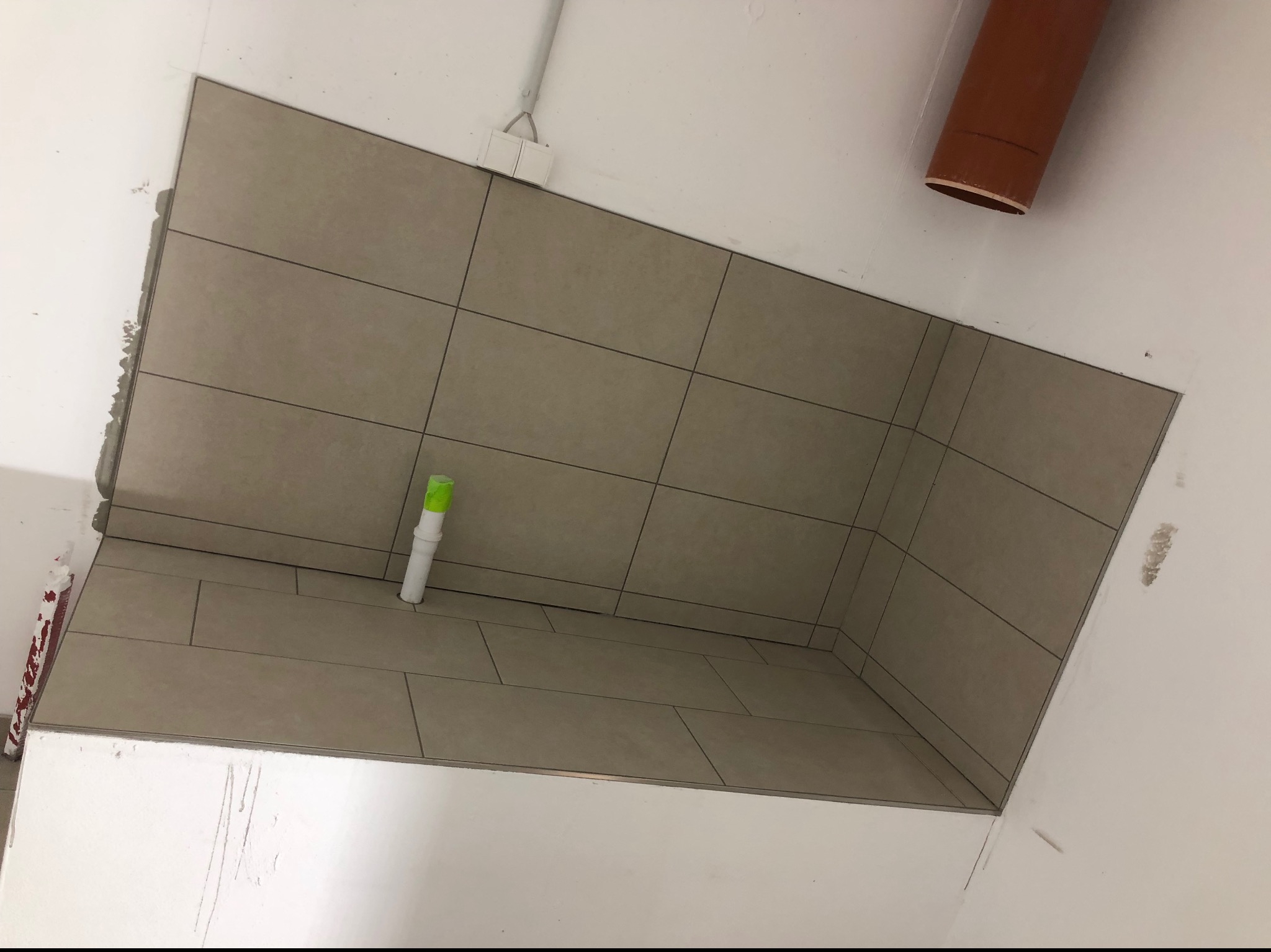 Kąt ujęcia na fragment wykończonej łazienki z beżowymi płytkami na ścianie i podłodze, widoczna rura kanalizacyjna z zielonym zabezpieczeniem oraz fragment brązowej rury na suficie.