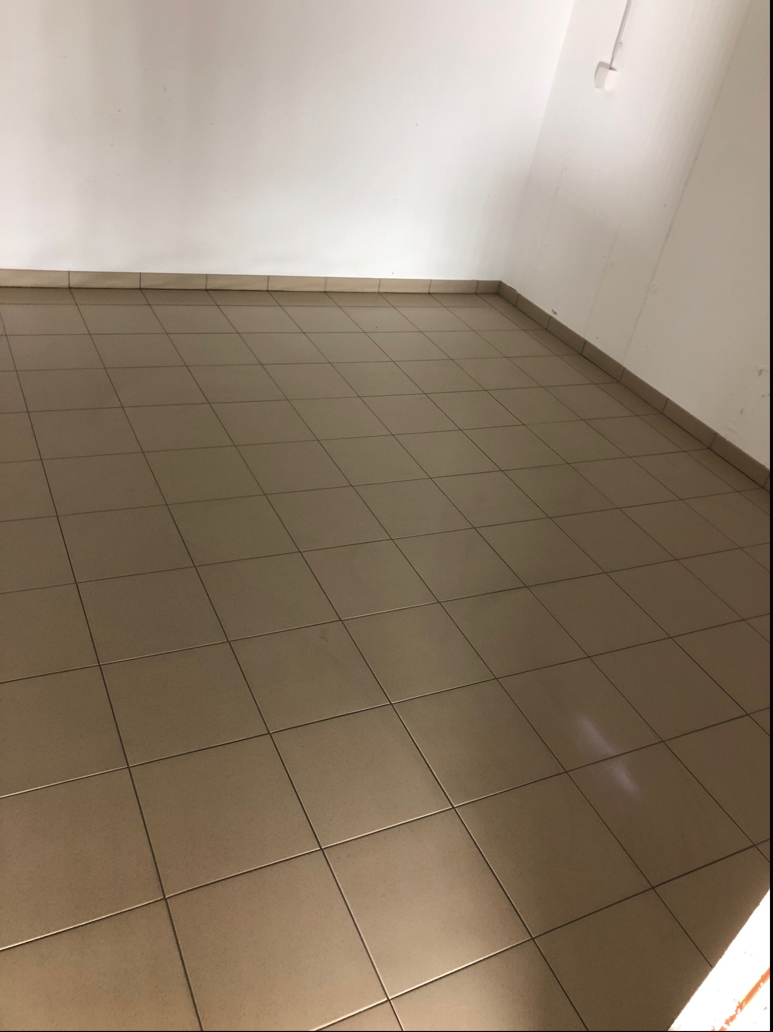 Beżowe, kwadratowe płytki podłogowe ułożone w regularną siatkę, widoczne przy białej ścianie z wystającym kablem elektrycznym.