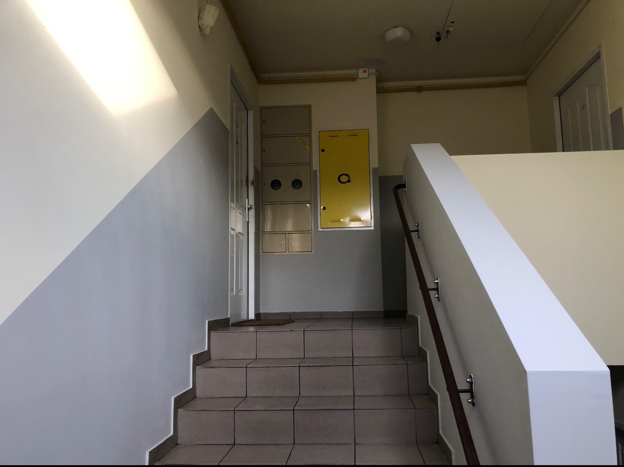 Klatka schodowa w bloku z szarymi schodami, ściany pomalowane do połowy na szaro, na ścianie szafki i żółta skrzynka z logo.