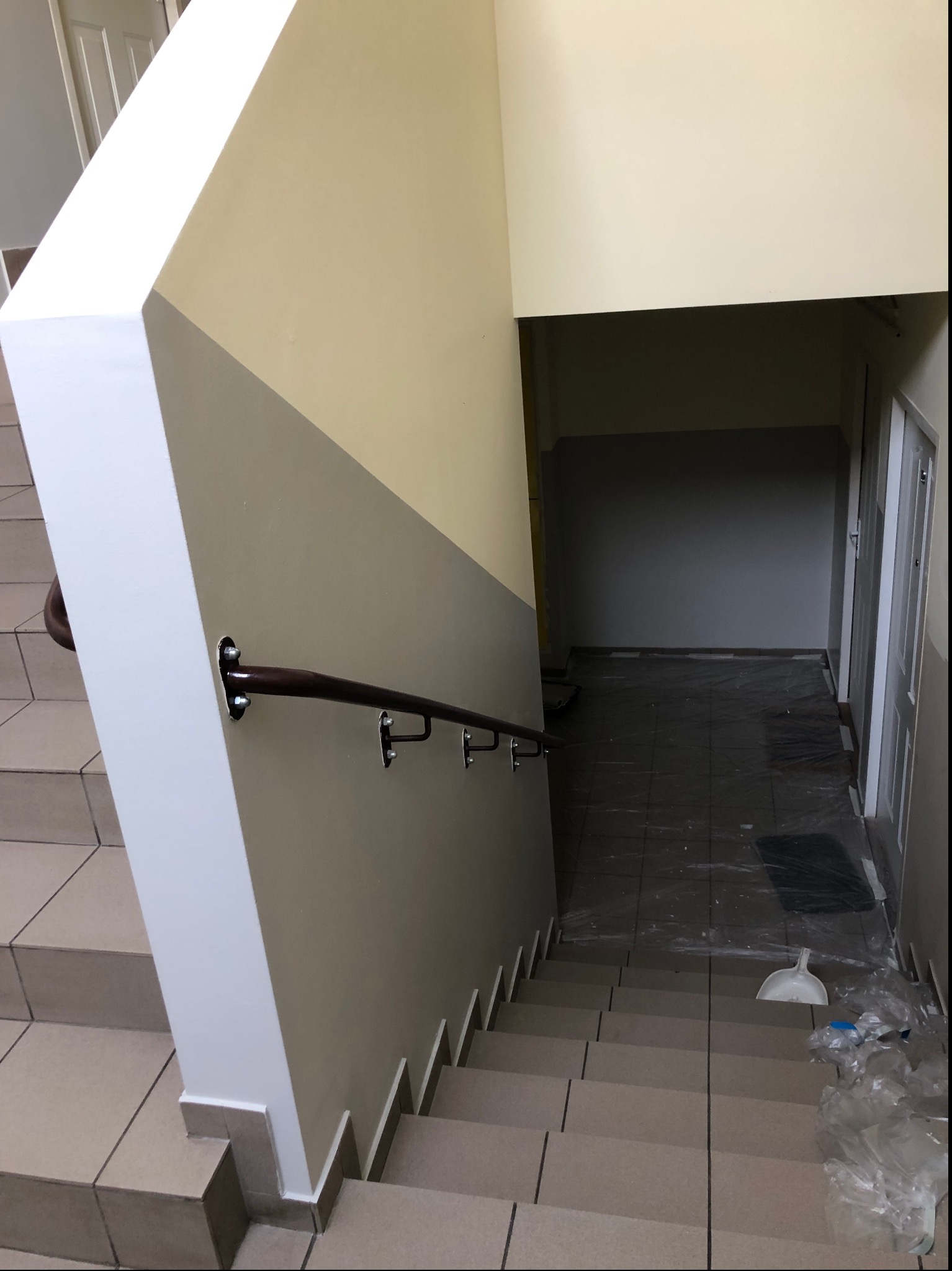 Klatka schodowa w trakcie prac wykończeniowych: pomalowane ściany w dwóch odcieniach, zabezpieczone folią podłogi i schody, widoczna poręcz i akcesoria do sprzątania.