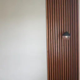 Panel ścienny z pionowych drewnianych listew w odcieniach brązu i czerni, oświetlony wiszącą lampą z przezroczystym kloszem, na tle szarej ściany.