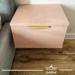 Różowa szafka nocna z szufladą i złotym uchwytem, stojąca na drewnianej podłodze obok szarej sofy.
