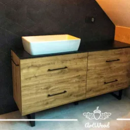 Drewniana szafka łazienkowa z czarnym blatem pod umywalką nablatową, czarne podłużne uchwyty, skośna ściana z ciemnymi płytkami w tle.