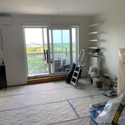 Yaroslav Pass Sp. z o.o. - Przygotowanie do malowania pokoju z balkonem: podłoga zabezpieczona papierem malarskim z taśmą, widoczne drabiny, farby, narzędzia i materiały budowlane, białe półki na ścianie.