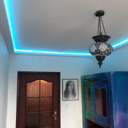 Yaroslav Pass Sp. z o.o. - Wnętrze z podświetlanym sufitem LED, mozaikową lampą wiszącą, ciemnymi drzwiami z szybą, portretem na ścianie i kolorową szafką w odcieniach niebieskiego i zieleni.