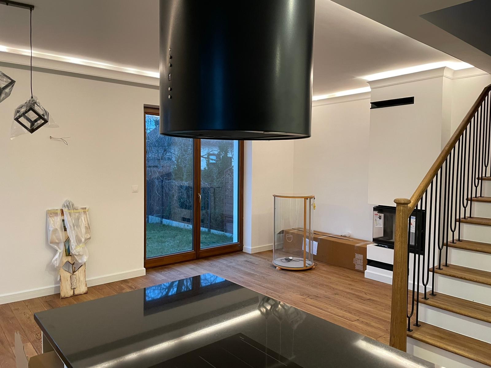 Wnętrze domu w Warszawie po remoncie: widok na drewniane schody z czarną balustradą, czarny okap nad wyspą kuchenną, przeszklone drzwi balkonowe, białe ściany z listwami LED pod sufitem...