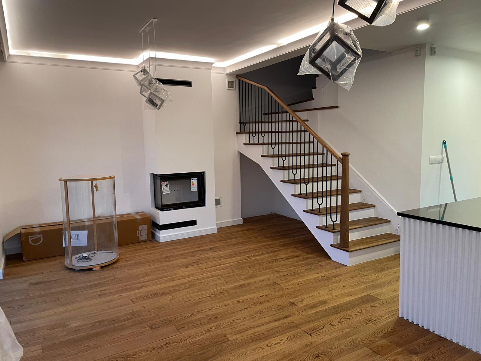 Salon z drewnianą podłogą, schodami z czarną balustradą i kominkiem, lampy wiszące w folii ochronnej, częściowo umeblowany.